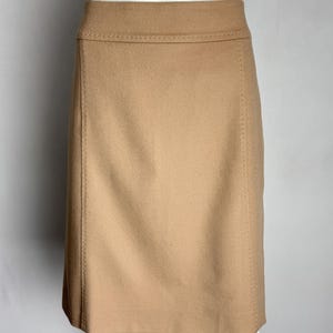 Puede incluir: Falda lápiz de color beige claro exhibida en un maniquí. La falda tiene una silueta recta, que llega hasta las rodillas, y presenta costuras visibles a lo largo de las costuras y la cintura. La tela parece ser lana suave o una mezcla de lana. Mide unos 60 cm de largo.