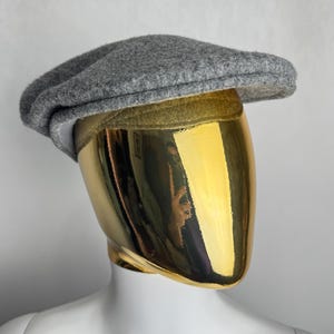 Pode incluir: Bon&eacute; cinzento, estilo newsboy, feito de mistura de l&atilde;, com uma coroa arredondada e uma aba pequena e r&iacute;gida. O chap&eacute;u &eacute; exibido em um manequim com cabe&ccedil;a dourada e torso branco.