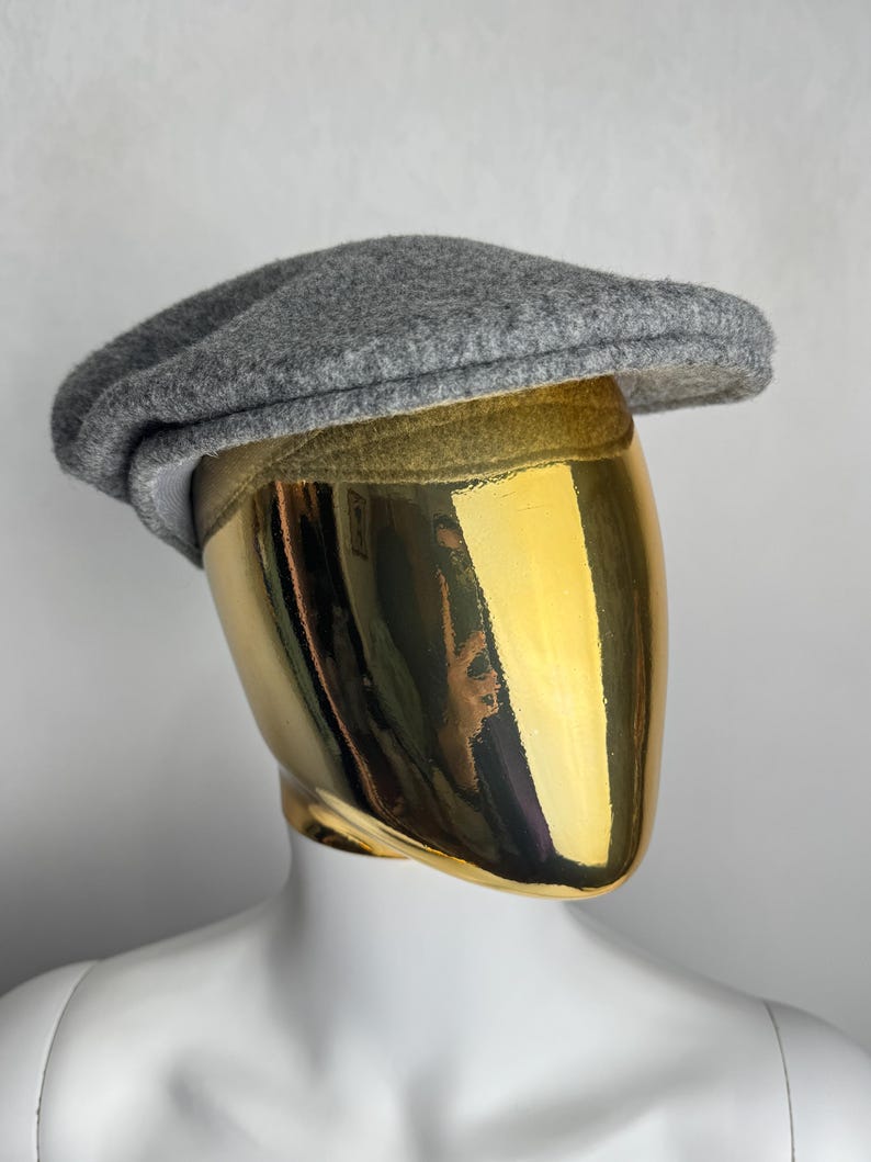 Pode incluir: Bon&eacute; cinzento, estilo newsboy, feito de mistura de l&atilde;, com uma coroa arredondada e uma aba pequena e r&iacute;gida. O chap&eacute;u &eacute; exibido em um manequim com cabe&ccedil;a dourada e torso branco.