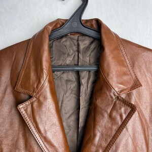 May include: A brown leather blazer hanging on a black hanger. The blazer has a classic design with a notched collar, two front pockets, and a button closure. The leather has a rich, textured appearance.