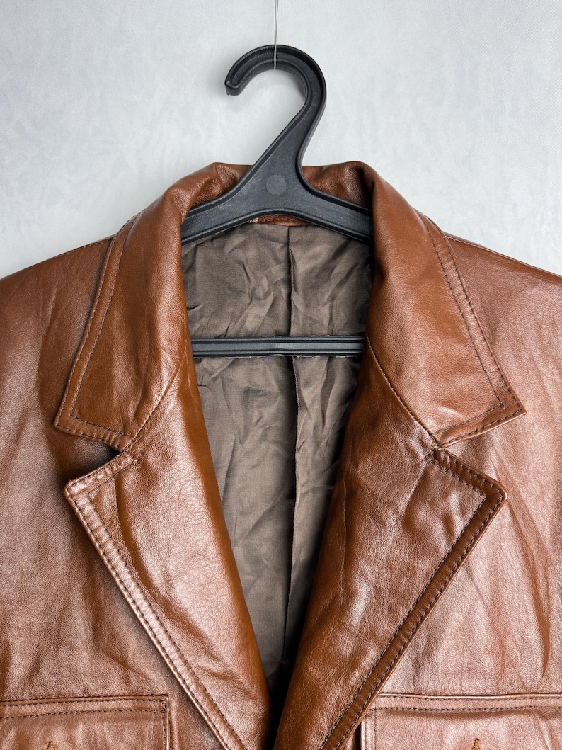 May include: A brown leather blazer hanging on a black hanger. The blazer has a classic design with a notched collar, two front pockets, and a button closure. The leather has a rich, textured appearance.