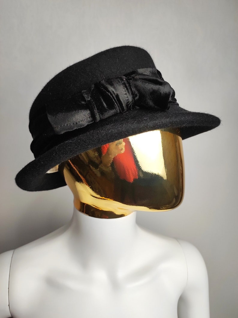 May include: A black cloche hat with a wide brim and a decorative satin bow. The hat is made of a felt-like material and is displayed on a mannequin head with a gold metallic finish. A classic accessory.