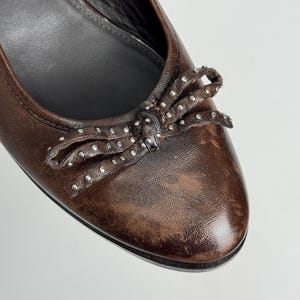 Puede incluir: Primer plano de una bailarina de cuero marr&oacute;n con un lazo adornado con peque&ntilde;os tachuelas plateadas. El zapato tiene un acabado envejecido y una puntera redondeada. La suela es negra.