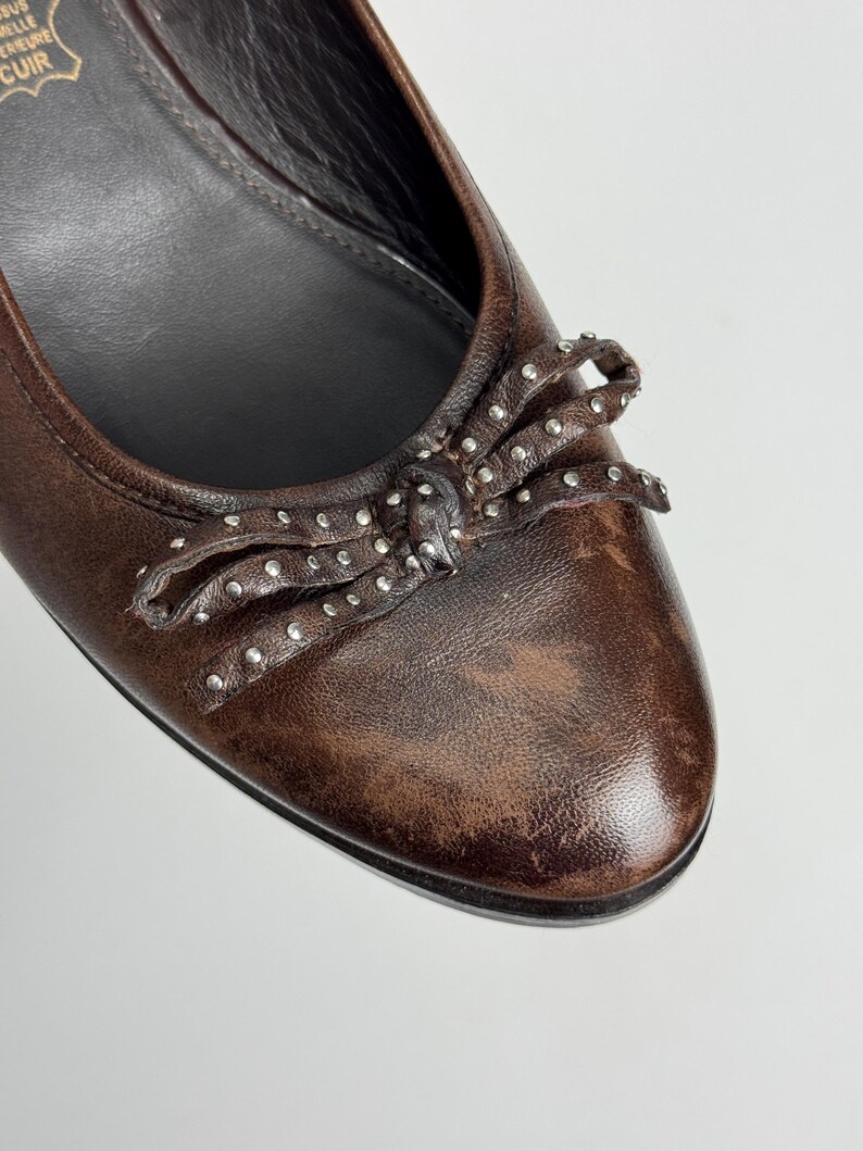 Puede incluir: Primer plano de una bailarina de cuero marr&oacute;n con un lazo adornado con peque&ntilde;os tachuelas plateadas. El zapato tiene un acabado envejecido y una puntera redondeada. La suela es negra.