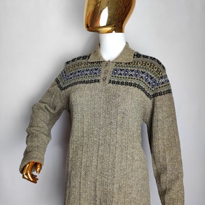 May include: A long-sleeved, collared sweater in a neutral beige color. The sweater features a patterned design across the shoulders in shades of blue, green, and black. The ribbed knit fabric adds texture to the garment.