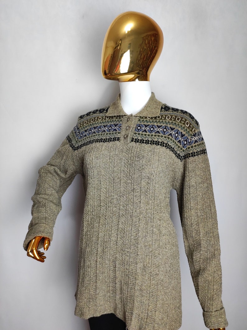 May include: A long-sleeved, collared sweater in a neutral beige color. The sweater features a patterned design across the shoulders in shades of blue, green, and black. The ribbed knit fabric adds texture to the garment.