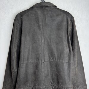 Puede incluir: Una chaqueta de cuero marr&oacute;n oscuro cuelga de una percha negra. La chaqueta tiene cuello, mangas largas y cierre de botones. La parte trasera de la chaqueta es visible, mostrando las costuras y los paneles del dise&ntilde;o.