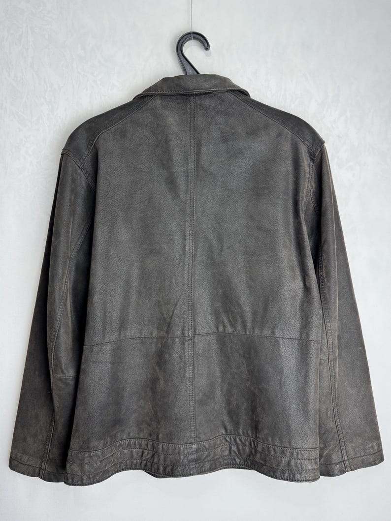 Puede incluir: Una chaqueta de cuero marr&oacute;n oscuro cuelga de una percha negra. La chaqueta tiene cuello, mangas largas y cierre de botones. La parte trasera de la chaqueta es visible, mostrando las costuras y los paneles del dise&ntilde;o.