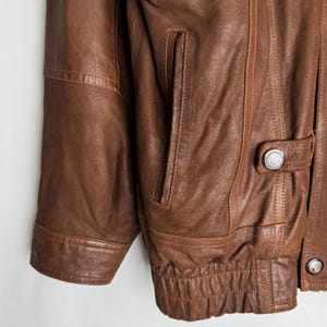 May include: A brown leather jacket with multiple pockets and button details. The jacket features a ribbed hem and cuffs, and a classic design. The leather appears to be well-worn, giving it a vintage look. The jacket has a classic, timeless style.