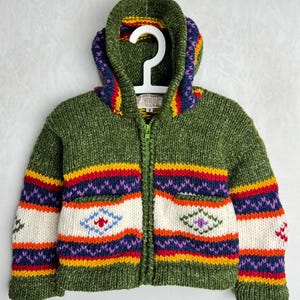 May include: A green hooded zip-up sweater with colourful striped accents. The sweater features a white central panel with embroidered geometric designs, and a green zip. The hood and cuffs have vibrant rainbow-coloured details.