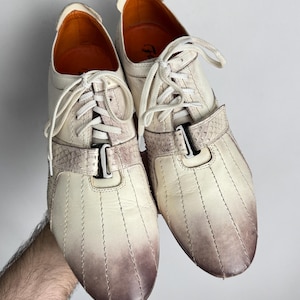 K&ouml;nnte beinhalten: Cremefarbene Ledersneaker mit braunem Farbverlauf an der Spitze. Die Schuhe haben wei&szlig;e Schn&uuml;rsenkel, einen dekorativen Riemen und eine Metallschnalle. Das Innenfutter ist orange.