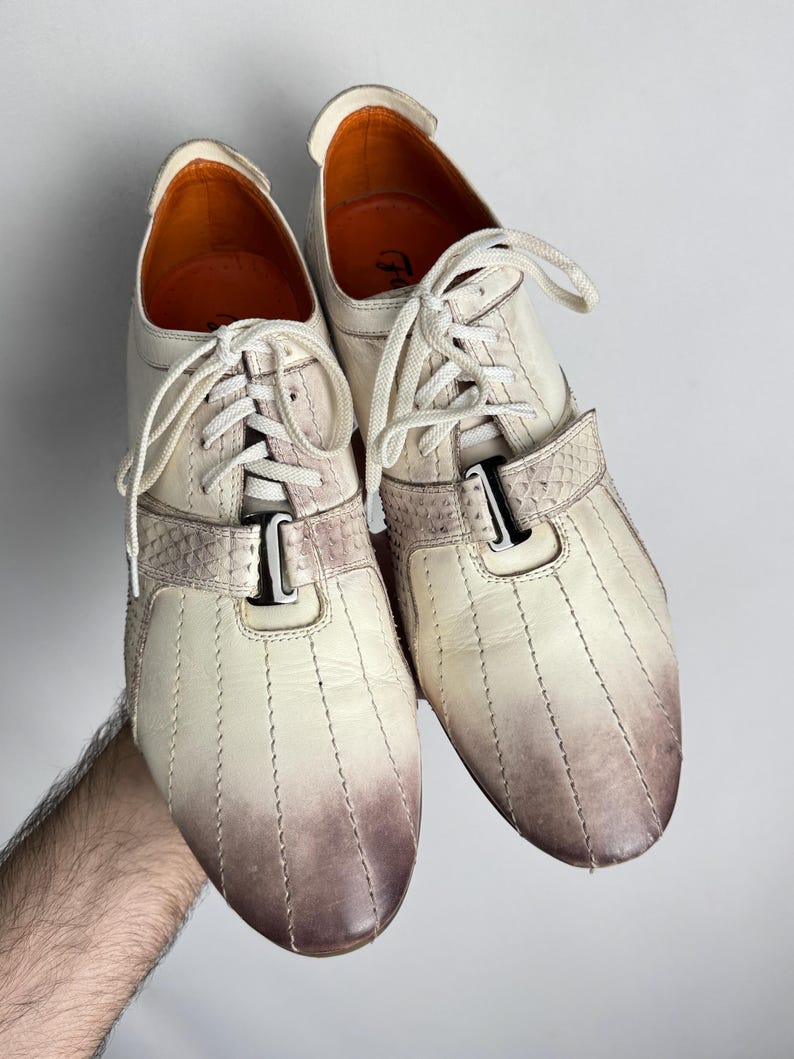 K&ouml;nnte beinhalten: Cremefarbene Ledersneaker mit braunem Farbverlauf an der Spitze. Die Schuhe haben wei&szlig;e Schn&uuml;rsenkel, einen dekorativen Riemen und eine Metallschnalle. Das Innenfutter ist orange.