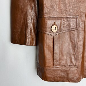 May include: A close-up of a brown leather jacket with a pocket and a button. The jacket has a classic design with a tailored fit. The leather appears to be soft and supple, with visible grain and texture.