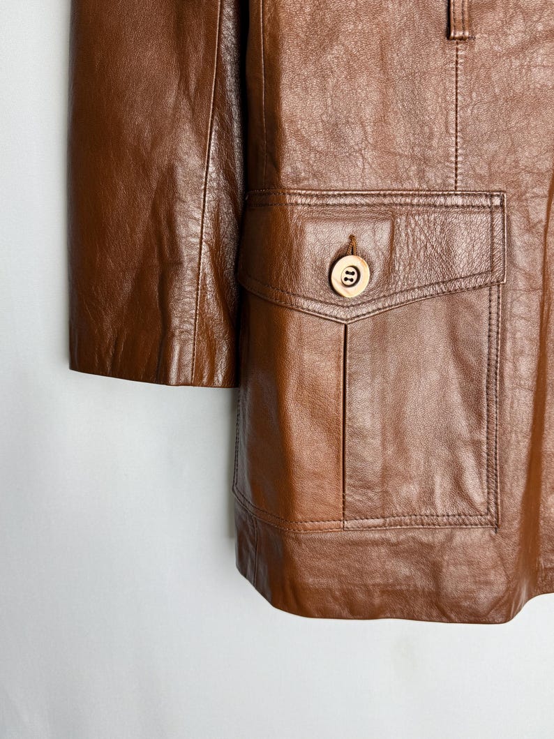May include: A close-up of a brown leather jacket with a pocket and a button. The jacket has a classic design with a tailored fit. The leather appears to be soft and supple, with visible grain and texture.