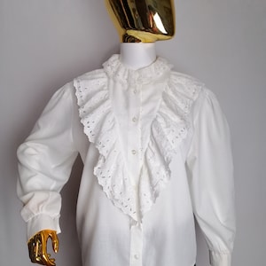 Puede incluir: Blusa blanca de manga larga con un gran cuello de volantes y detalles de encaje de ojal.