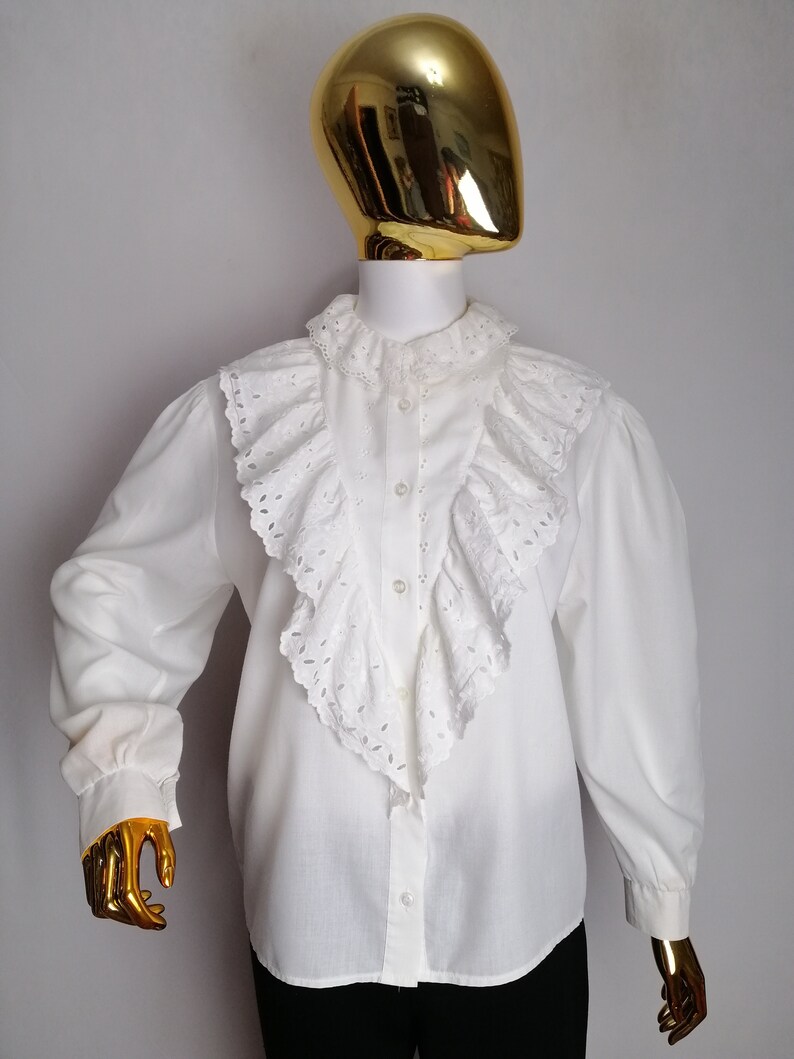 Puede incluir: Blusa blanca de manga larga con un gran cuello de volantes y detalles de encaje de ojal.