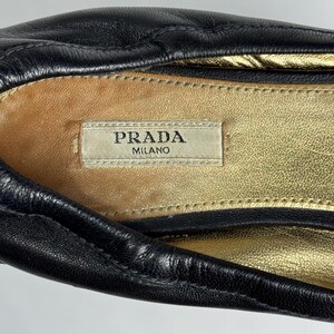 Puede incluir: Primer plano de un zapato de cuero negro con una plantilla dorada. El interior del zapato presenta una etiqueta que dice "PRADA MILANO". El cuero parece suave y texturizado.