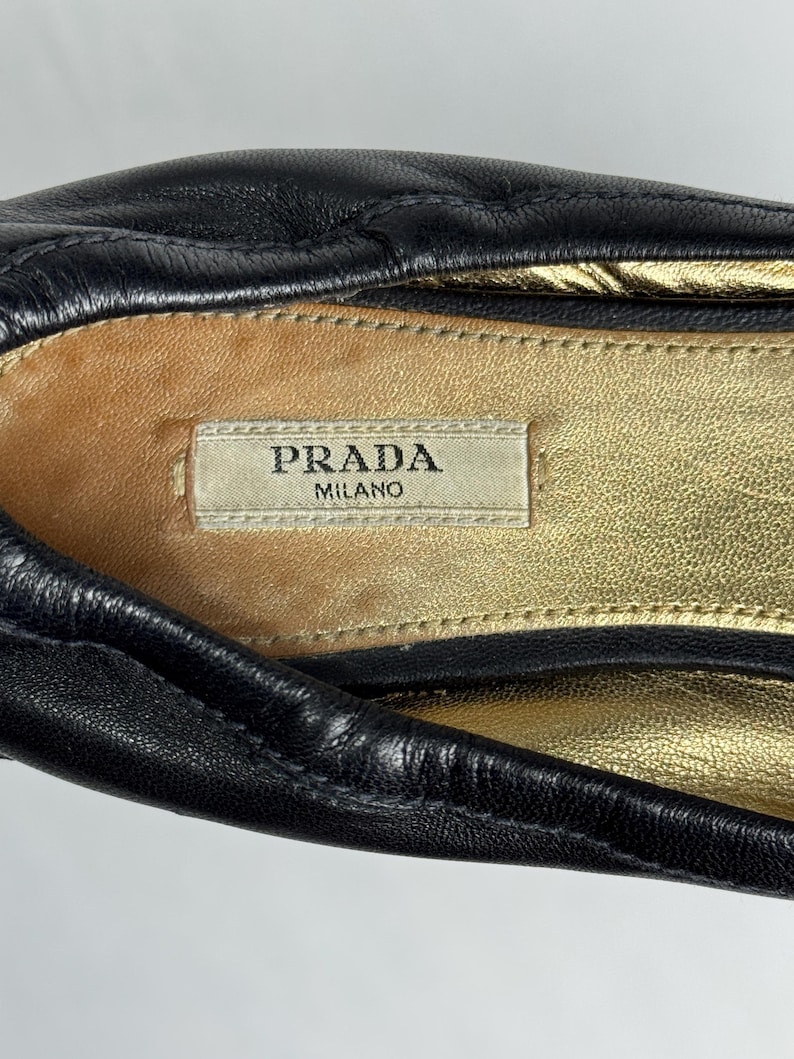 Puede incluir: Primer plano de un zapato de cuero negro con una plantilla dorada. El interior del zapato presenta una etiqueta que dice "PRADA MILANO". El cuero parece suave y texturizado.