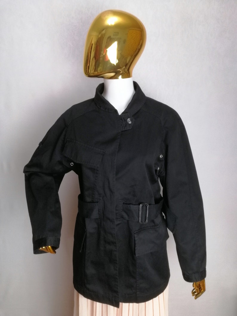 May include: A black utility jacket with a belted waist, multiple pockets, and a stand-up collar.