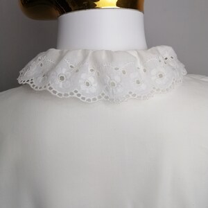 Puede incluir: Una blusa blanca con un cuello de encaje blanco. El cuello tiene un borde festoneado y un delicado patr&oacute;n floral.