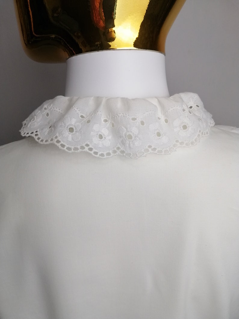 Puede incluir: Una blusa blanca con un cuello de encaje blanco. El cuello tiene un borde festoneado y un delicado patr&oacute;n floral.