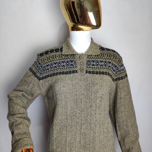 May include: A long-sleeved, collared sweater in a neutral beige color. The sweater features a patterned yoke with blue, green, and black geometric designs. The garment is displayed on a mannequin with a gold head.