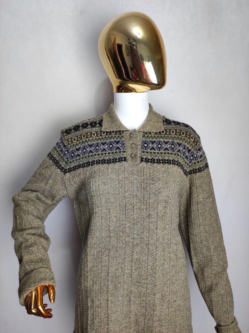 May include: A long-sleeved, collared sweater in a neutral beige color. The sweater features a patterned yoke with blue, green, and black geometric designs. The garment is displayed on a mannequin with a gold head.
