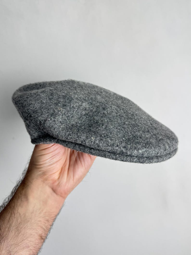 Pode incluir: Bon&eacute; plano cinzento de l&atilde;. O chap&eacute;u tem uma coroa arredondada e uma aba pequena e r&iacute;gida. O bon&eacute; &eacute; segurado contra um fundo branco.