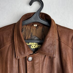 May include: A brown leather jacket with a button closure and a collar, hanging on a black hanger. The jacket has the text "CAVAL CONTI" in gold lettering. The jacket is a medium size.