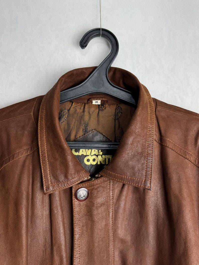 May include: A brown leather jacket with a button closure and a collar, hanging on a black hanger. The jacket has the text "CAVAL CONTI" in gold lettering. The jacket is a medium size.
