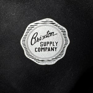 Puede incluir: Una etiqueta circular blanca con texto negro que dice "Brixton Supply Company" sobre un fondo negro.