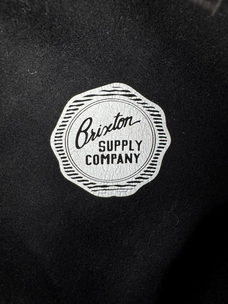 Puede incluir: Una etiqueta circular blanca con texto negro que dice "Brixton Supply Company" sobre un fondo negro.