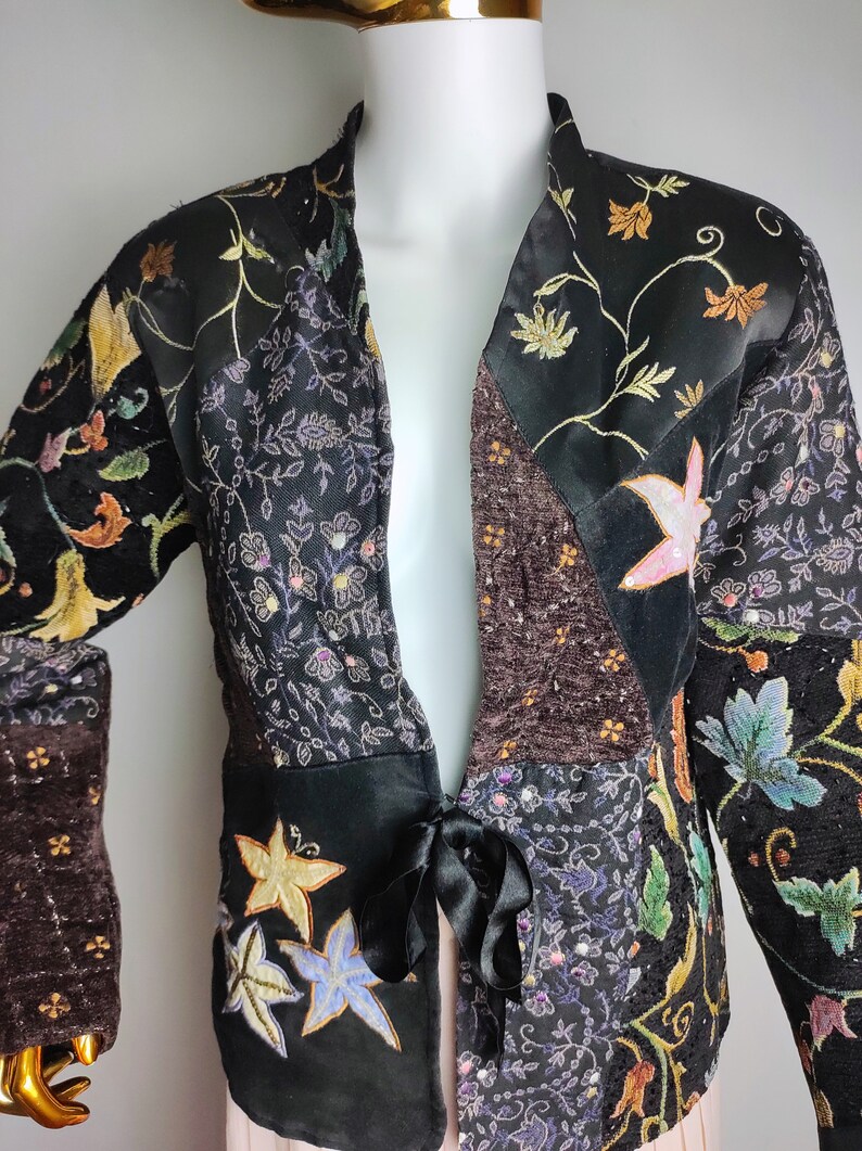 May include: A black jacket with a floral pattern and a tie at the waist. The jacket is made of different fabrics and has a patchwork design.