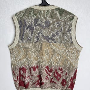 May include: A sleeveless sweater vest with a patterned design in shades of beige, green, gray, and burgundy. The vest is hanging on a black hanger against a white background. The knitwear has a ribbed hem and neckline.