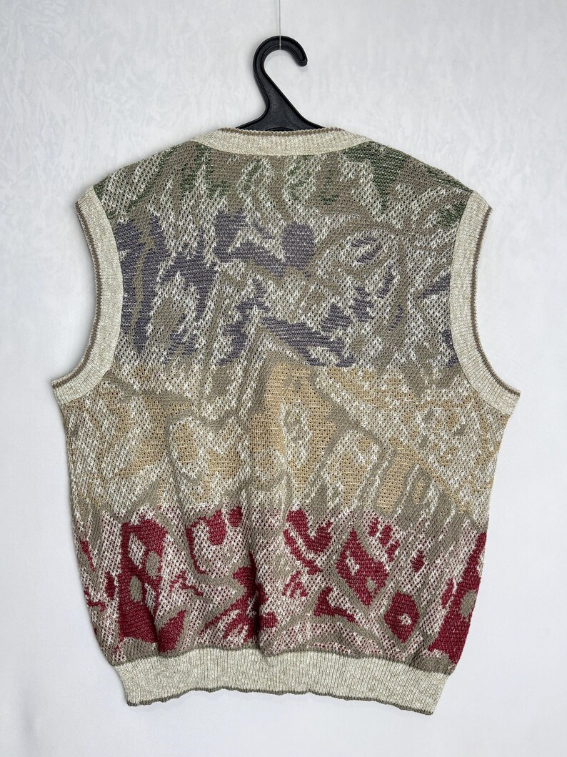 May include: A sleeveless sweater vest with a patterned design in shades of beige, green, gray, and burgundy. The vest is hanging on a black hanger against a white background. The knitwear has a ribbed hem and neckline.