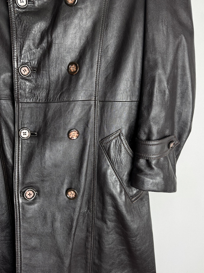 May include: A dark brown leather coat with a double-breasted design. The coat features multiple buttons, a notched collar, and a tailored fit. The coat has a classic, vintage style, with a pocket on the front.