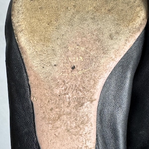 Puede incluir: Primer plano de la suela de un zapato de cuero negro. La suela est&aacute; desgastada y muestra el nombre de la marca "PRADA", la talla "37 1/2" y "MADE IN ITALY" en relieve sobre el cuero beige desgastado. Se ve la parte superior de cuero negro del zapato.