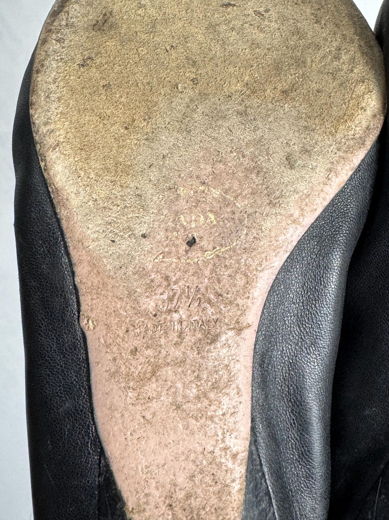 Puede incluir: Primer plano de la suela de un zapato de cuero negro. La suela est&aacute; desgastada y muestra el nombre de la marca "PRADA", la talla "37 1/2" y "MADE IN ITALY" en relieve sobre el cuero beige desgastado. Se ve la parte superior de cuero negro del zapato.