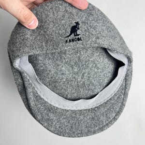 Pode incluir: Bon&eacute; plano Kangol cinzento com um log&oacute;tipo preto de um canguru e o nome da marca. O bon&eacute; &eacute; feito de um material macio semelhante a feltro e tem um forro interior cinzento. O bon&eacute; &eacute; segurado por uma m&atilde;o.