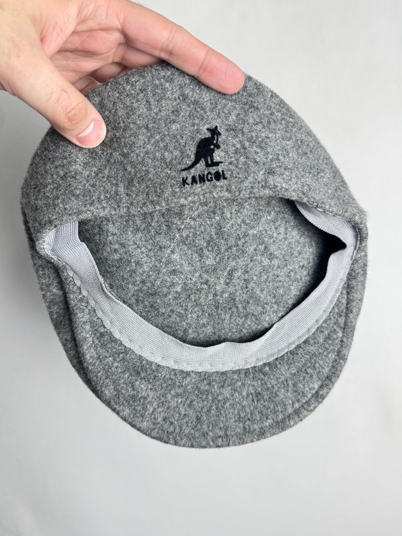 Pode incluir: Bon&eacute; plano Kangol cinzento com um log&oacute;tipo preto de um canguru e o nome da marca. O bon&eacute; &eacute; feito de um material macio semelhante a feltro e tem um forro interior cinzento. O bon&eacute; &eacute; segurado por uma m&atilde;o.