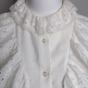 Puede incluir: Blusa blanca de encaje con ojales, con cuello de volantes y cierre de botones. La blusa tiene un delicado patr&oacute;n floral y es perfecta para una boda de verano o una ocasi&oacute;n especial.