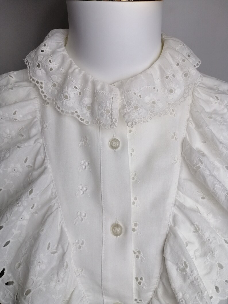 Puede incluir: Blusa blanca de encaje con ojales, con cuello de volantes y cierre de botones. La blusa tiene un delicado patr&oacute;n floral y es perfecta para una boda de verano o una ocasi&oacute;n especial.