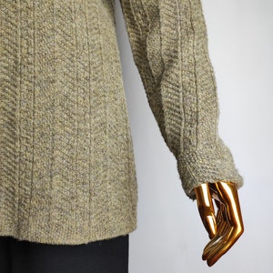May include: A textured, olive green sweater with a ribbed design on the sleeves and a subtle herringbone pattern on the body. The sweater has a crew neck and long sleeves. The bottom portion of the image shows black pants.