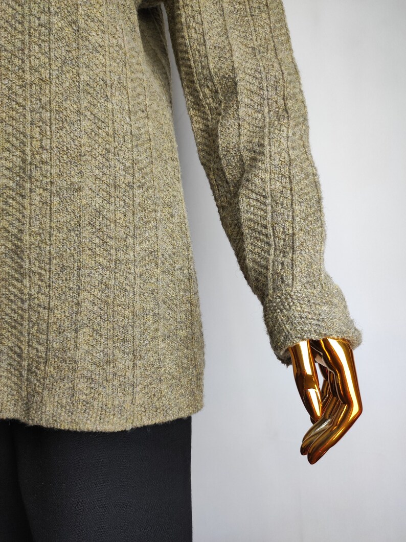 May include: A textured, olive green sweater with a ribbed design on the sleeves and a subtle herringbone pattern on the body. The sweater has a crew neck and long sleeves. The bottom portion of the image shows black pants.