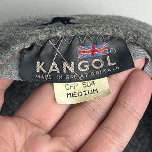 Pode incluir: Bon&eacute; Kangol cinzento com uma etiqueta preta que diz "KANGOL MADE IN GREAT BRITAIN" e a bandeira brit&acirc;nica. A etiqueta tamb&eacute;m mostra "CAP 504 MEDIUM."