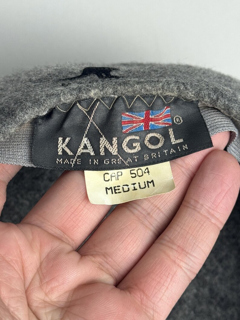 Pode incluir: Bon&eacute; Kangol cinzento com uma etiqueta preta que diz "KANGOL MADE IN GREAT BRITAIN" e a bandeira brit&acirc;nica. A etiqueta tamb&eacute;m mostra "CAP 504 MEDIUM."