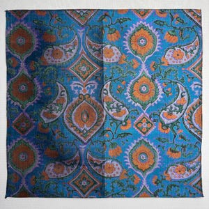 May include: A square textile with a vibrant paisley pattern. The design features large, stylized paisley shapes in shades of orange, green, and purple against a teal blue background. The fabric has a slightly textured appearance.