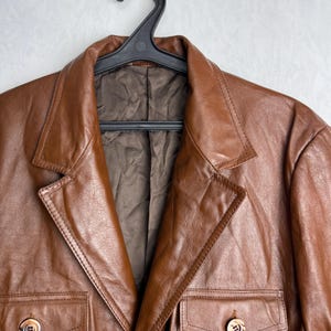 May include: A brown leather jacket with a classic design, featuring a notched collar, two chest pockets, and two lower pockets. The jacket is hanging on a black hanger. The leather has a smooth texture and a warm, rich color.