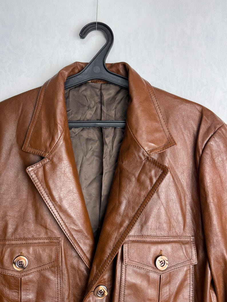 May include: A brown leather jacket with a classic design, featuring a notched collar, two chest pockets, and two lower pockets. The jacket is hanging on a black hanger. The leather has a smooth texture and a warm, rich color.