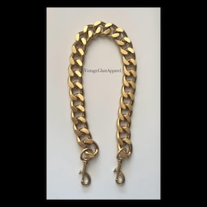 May include: A gold chain handbag strap with two clasps. The chain is thick and has a curb link design.